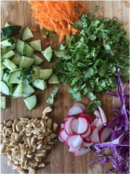 Chopped cucumbers, carrots, herbs, radishes, cabbage, and peanuts spread across a prep board